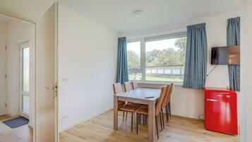 Une pièce comprenant une table à manger en bois, des chaises, un mini-réfrigérateur rouge, un téléviseur fixé au mur et une fenêtre avec des rideaux.