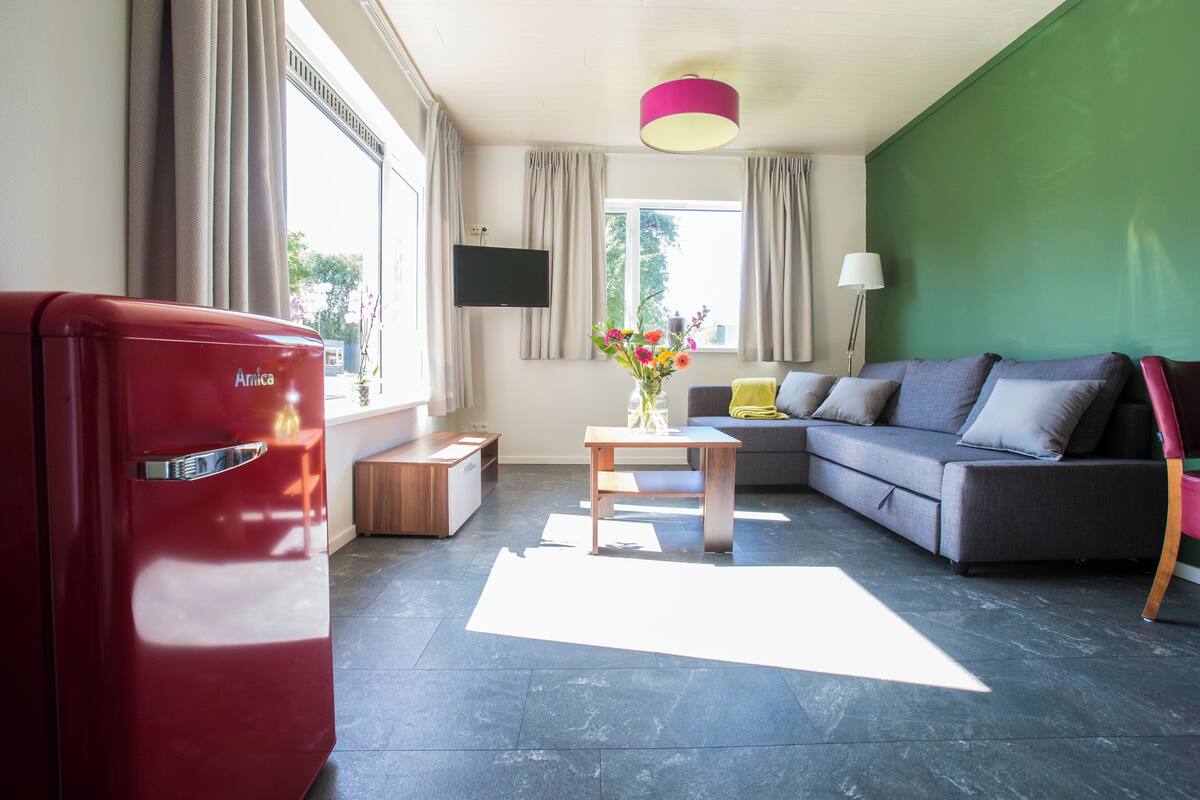 Una sala de estar moderna con un refrigerador rojo, un sofá gris, una mesa de centro de madera con flores y un televisor de pantalla plana montado en la pared.