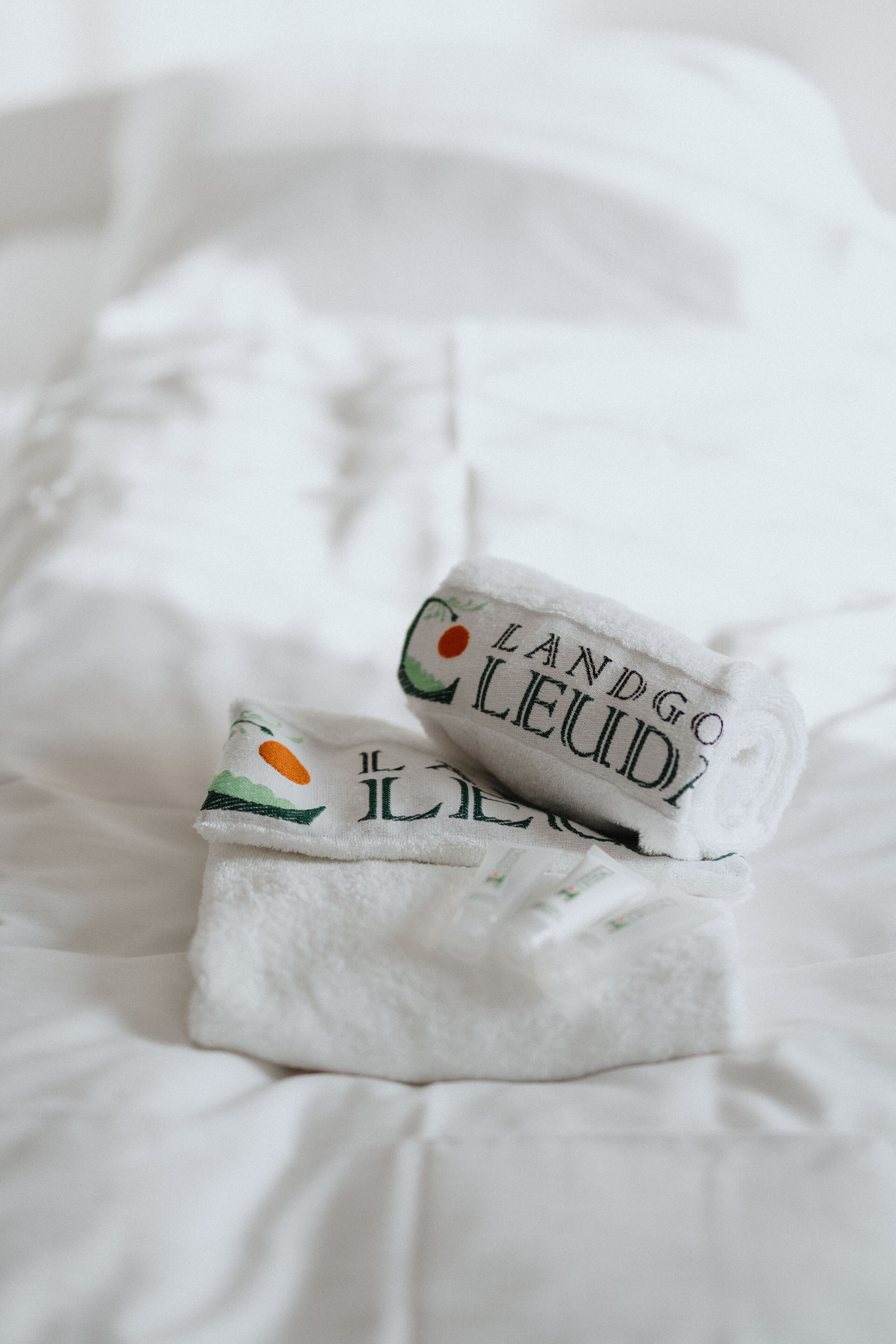 A neatly folded white towel with embroidered text and a small logo on a white bedspread.