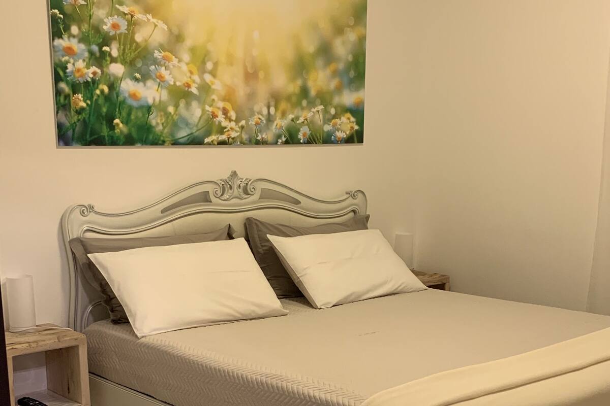 A neatly made bed with a white headboard and pillows, a bedside table with a lamp, and a painting of flowers on the wall.