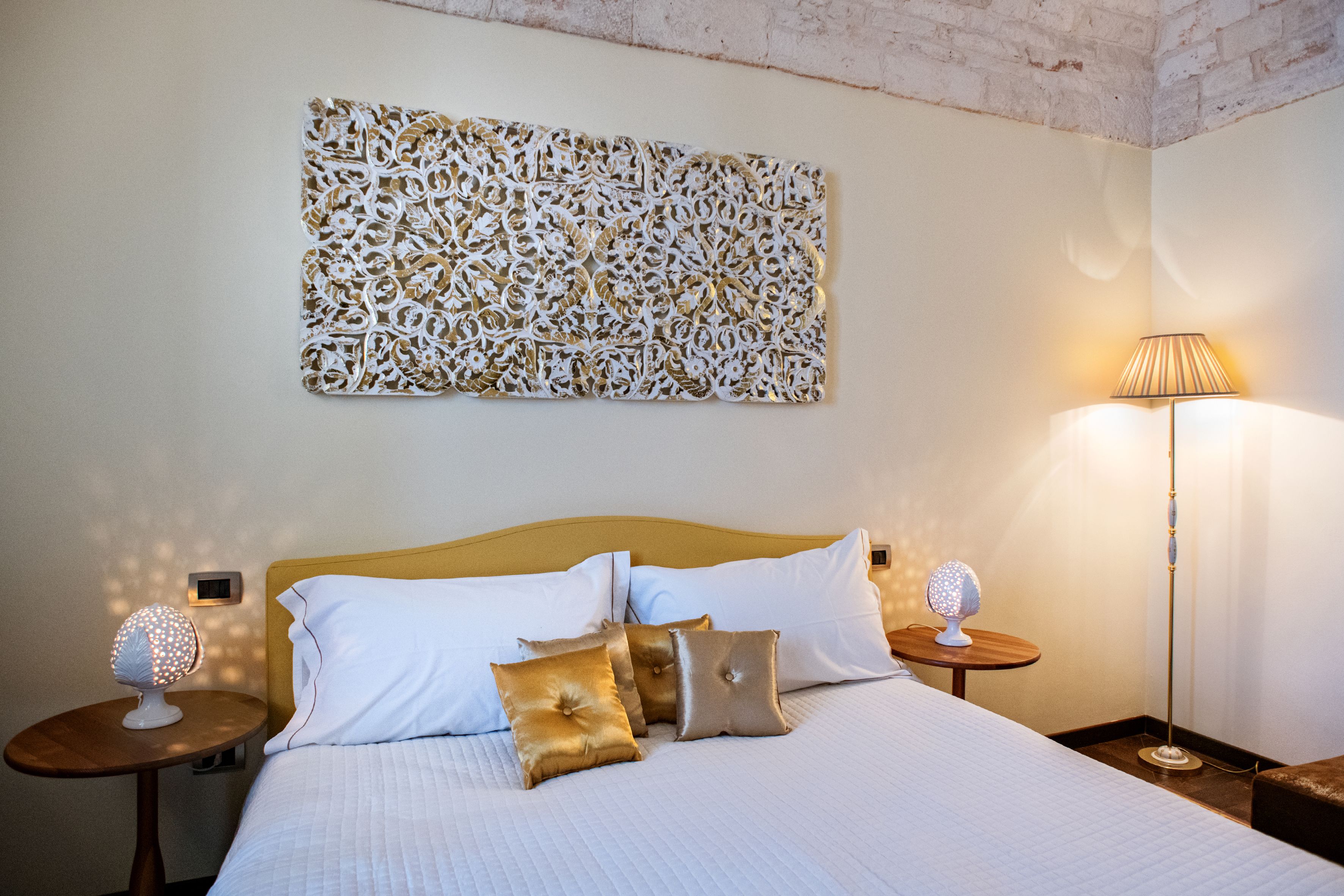 Una cama bien tendida con almohadas blancas y doradas, un adorno en la pared y dos mesitas de noche con lámparas.