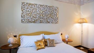 Una cama bien tendida con almohadas blancas y doradas, un adorno en la pared y dos mesitas de noche con lámparas.