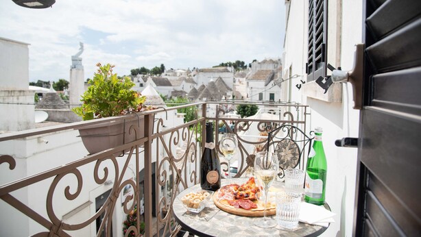 A balcony with a table set for a meal, including a bottle of wine, glasses, and a plate of food. - Perlage Suite Luxury B&B (Alberobello)