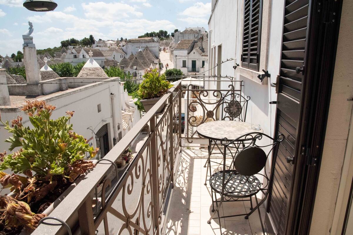 A balcony with a table and chairs, potted plants, and a view of a town with white buildings and a statue.