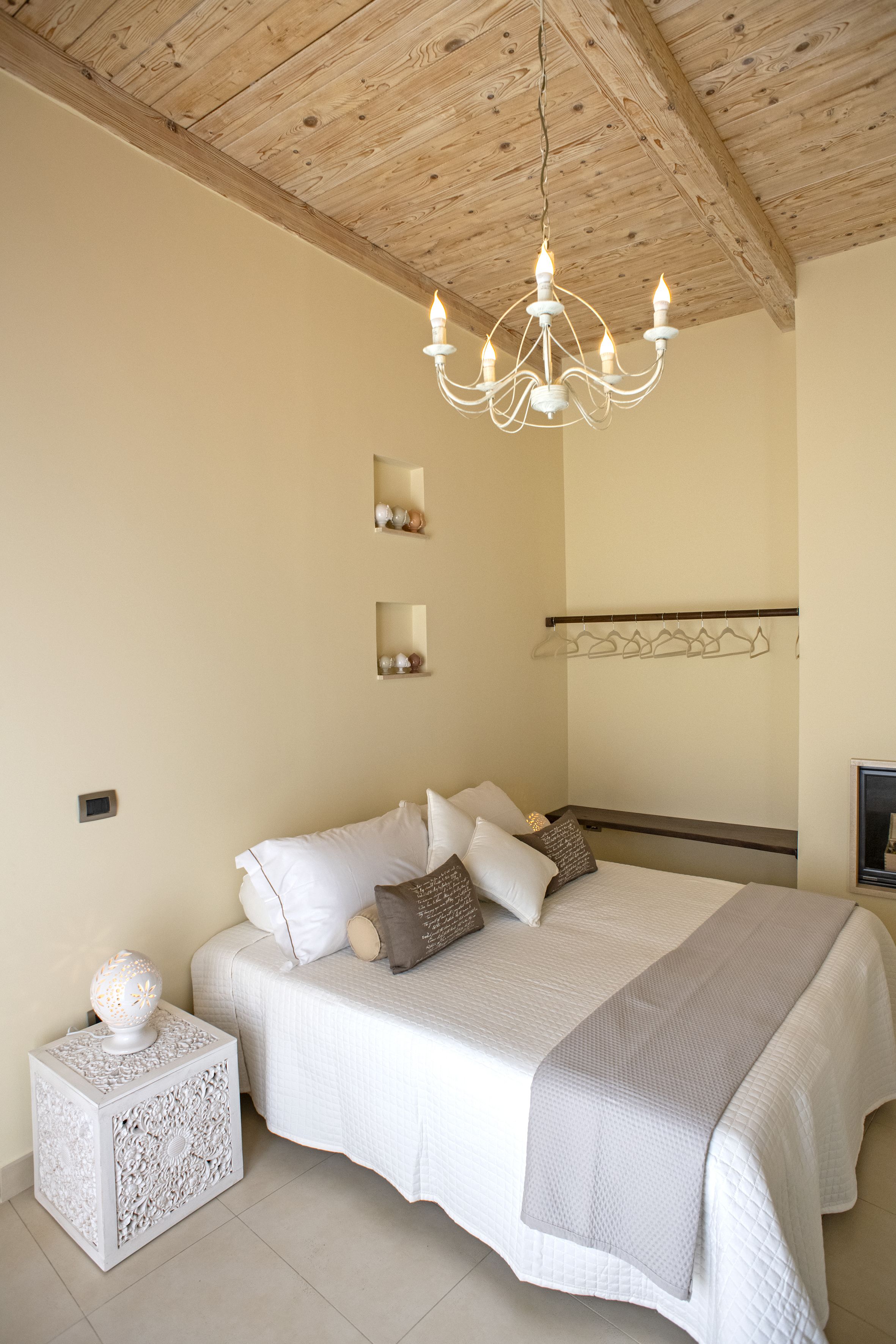 A bedroom with a wooden ceiling, a white bed with multiple pillows, a bedside table with a decorative item, and a wall-mounted mirror.