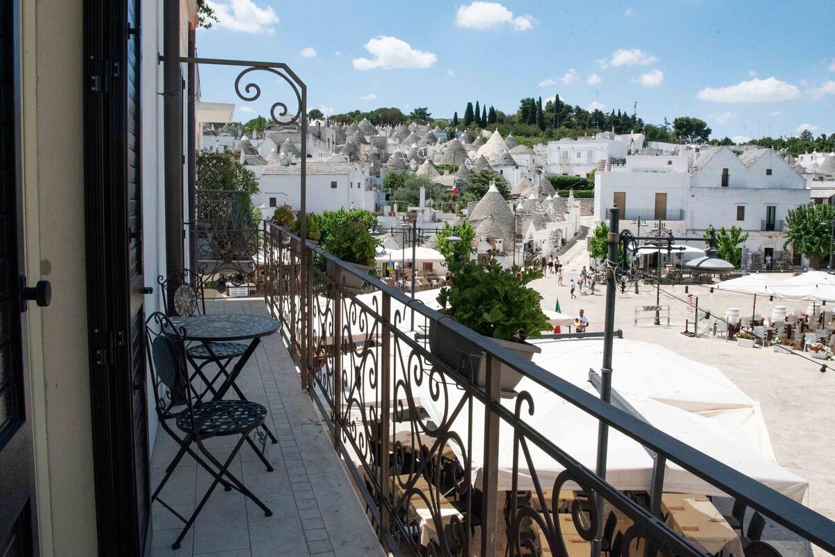 A balcony with a table and chairs, overlooking a town with white buildings and a clear sky.