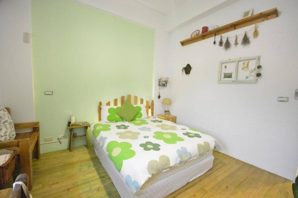 A bedroom with a bed, wooden headboard, bedside tables, a nightstand, a wall-mounted shelf with decorative items, and a wall clock.