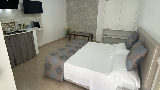 A bedroom with a bed, bedside table, and a small table with a vase. There is a kitchenette with a stove and a flat-screen TV mounted on the wall. - Casa FioRita (Vieste)