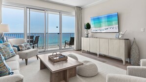 Un vaste salon doté d’une grande porte-fenêtre coulissante donnant sur un balcon avec vue sur l’océan. Un téléviseur à écran plat est fixé au mur, ainsi qu’une table basse en bois et un meuble blanc agrémenté d’objets décoratifs.
