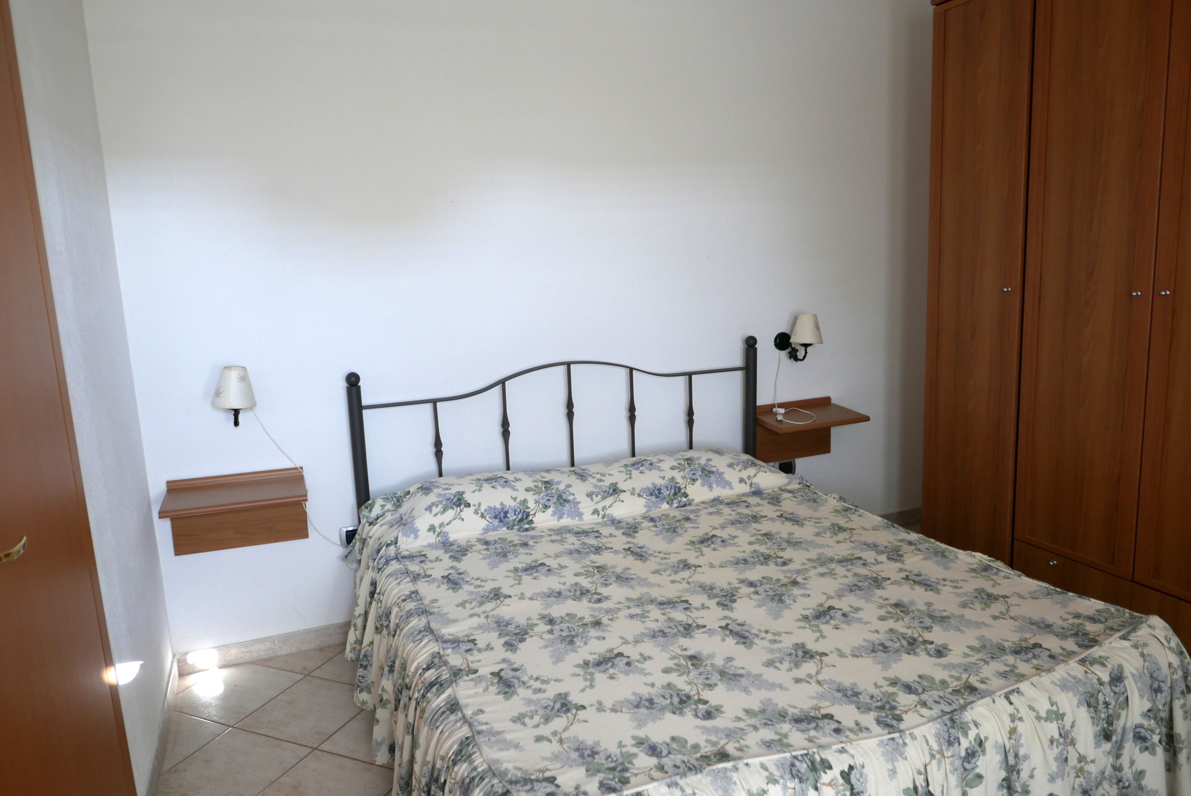 A bedroom with a bed, a nightstand, a wall-mounted lamp, a wooden wardrobe, and a small shelf.