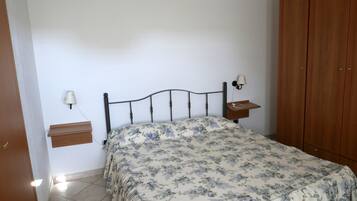 A bedroom with a bed, a nightstand, a wall-mounted lamp, a wooden wardrobe, and a small shelf.