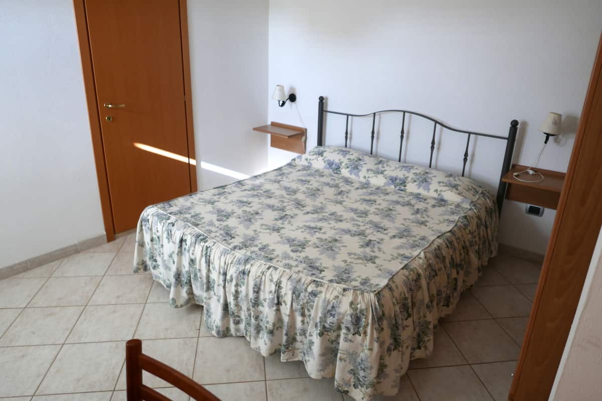 A bedroom with a bed, a wooden door, a nightstand, and a wall-mounted lamp.