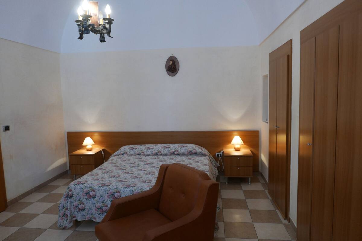 A bedroom with a bed, two bedside lamps, a wooden wardrobe, and a chandelier.