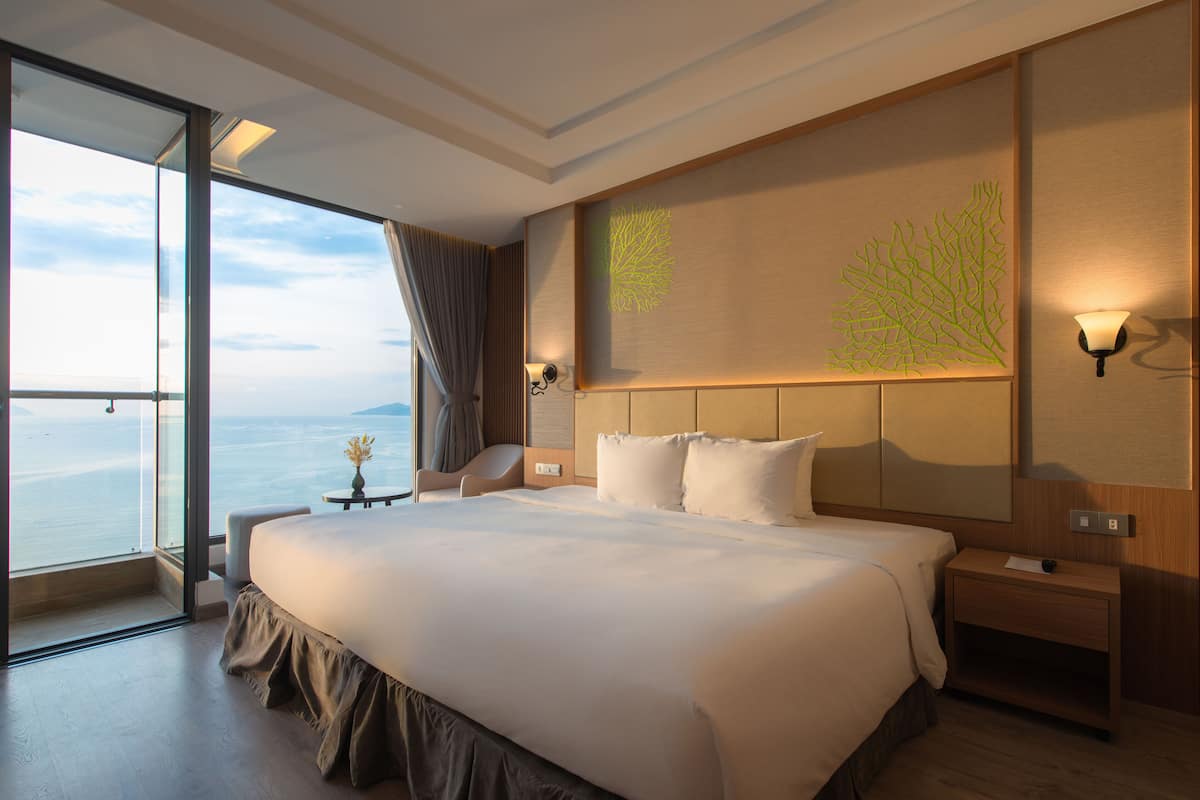 A modern hotel room with a large bed, a nightstand, a wall-mounted lamp, and a view of the sea through a large window.