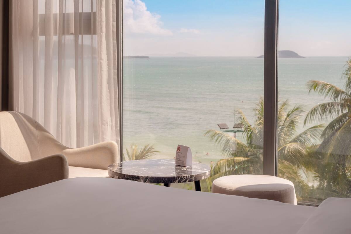 A hotel room with a large window overlooking the sea, a bed, a round table, and a chair.