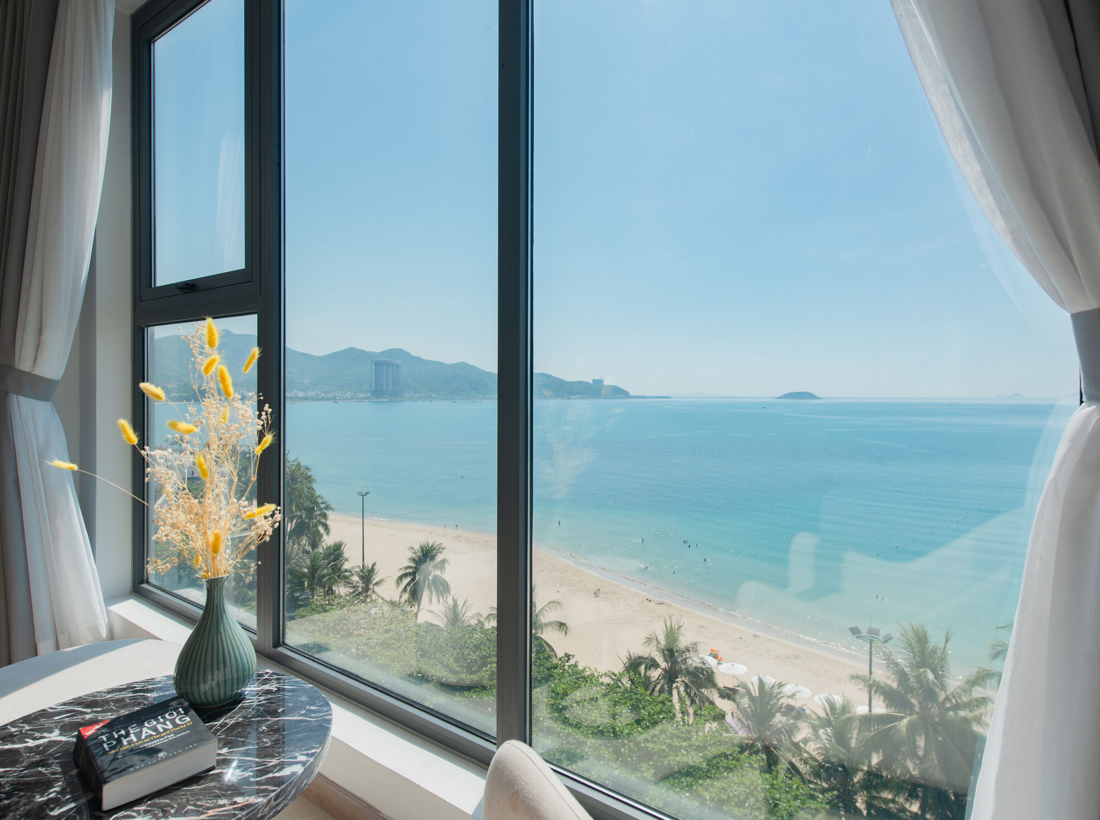 A room with a large window overlooking a beach, a vase with yellow flowers, and a book on a table.