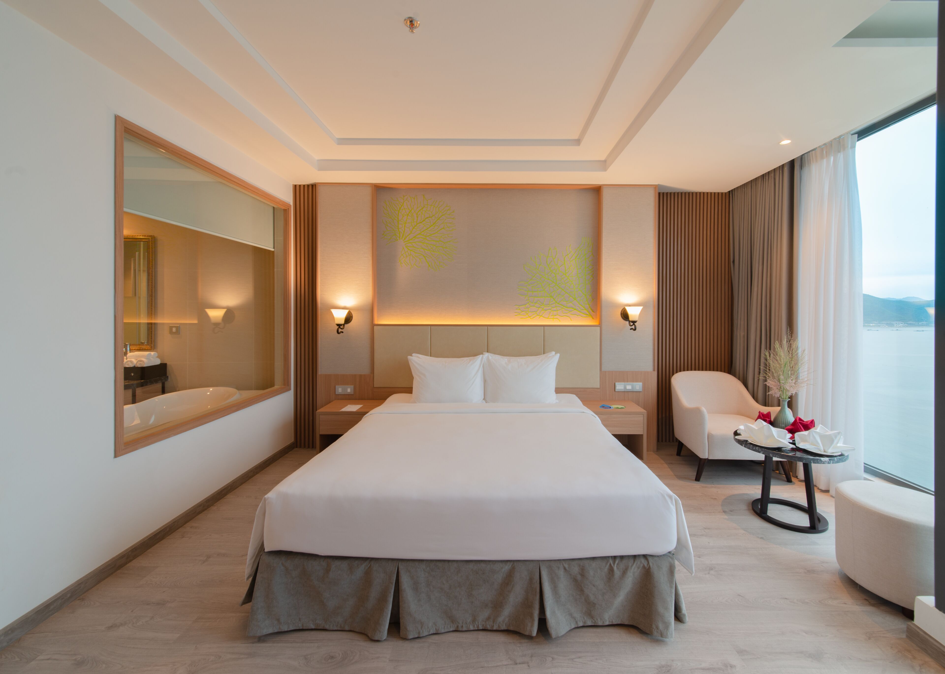 A modern hotel room with a large bed, a sitting area, a view of the sea, and a bathroom with a mirror and toiletries.