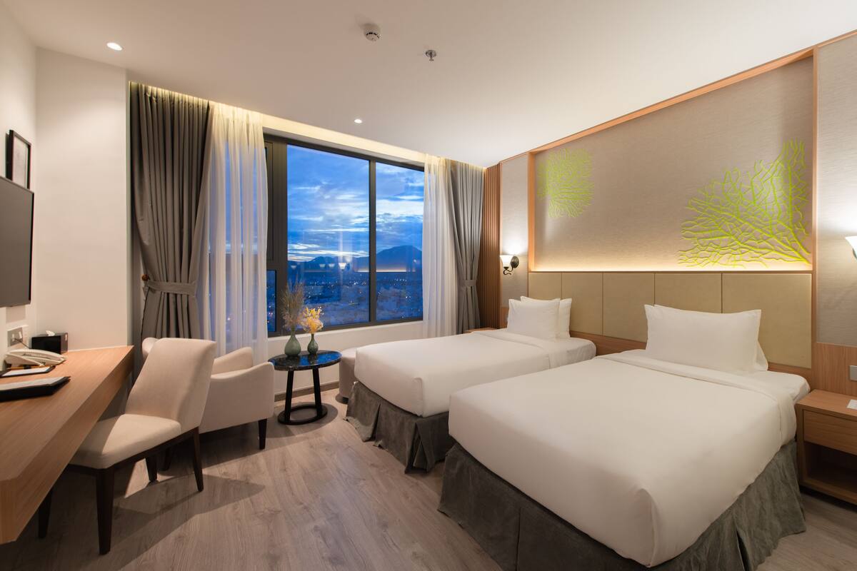 A hotel room with two beds, a desk, a chair, a small table with a vase, and a large window with curtains.