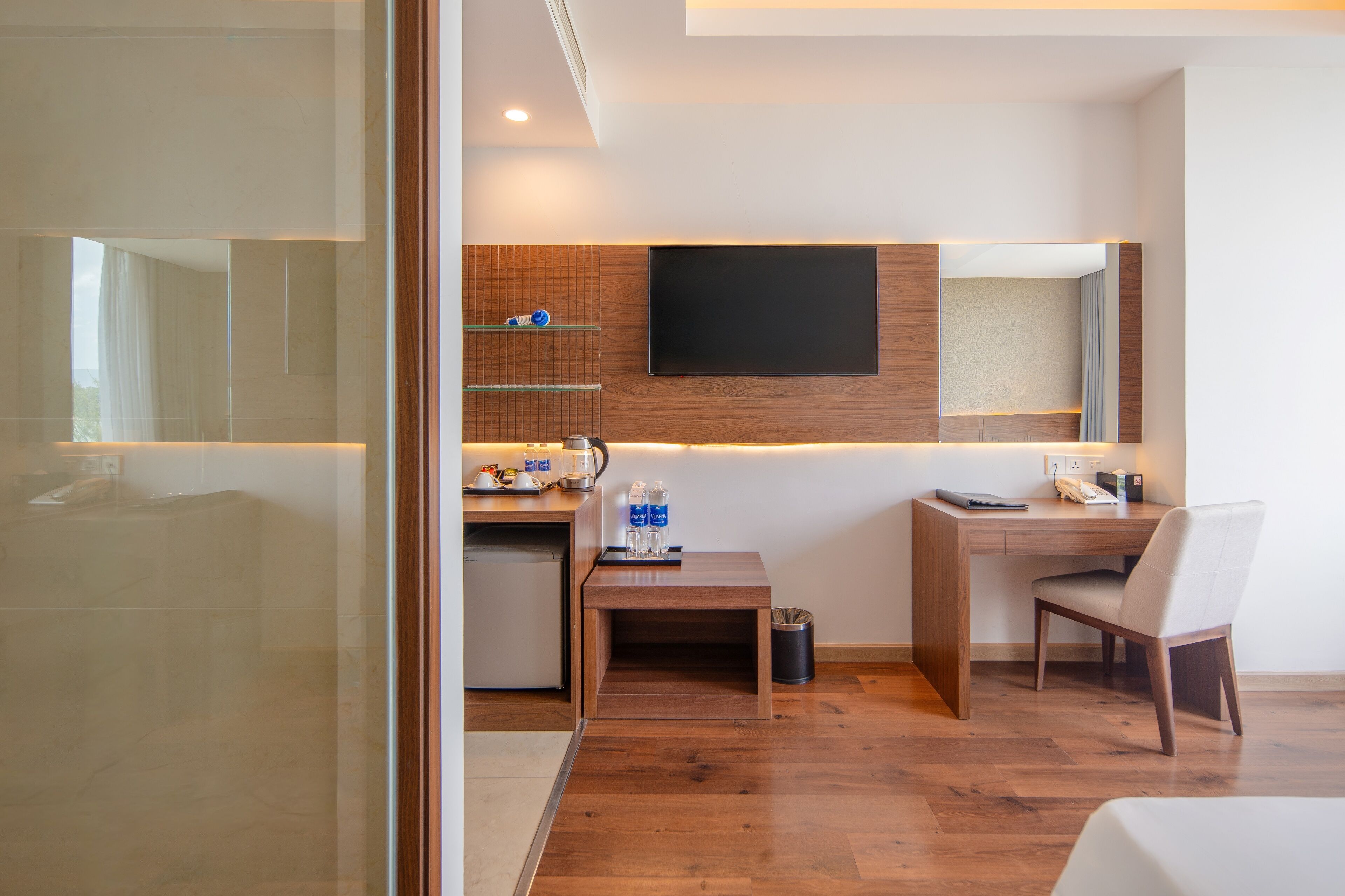 A modern hotel room with a wooden desk, a flat-screen TV mounted on the wall, a small kitchenette, and a large mirror.