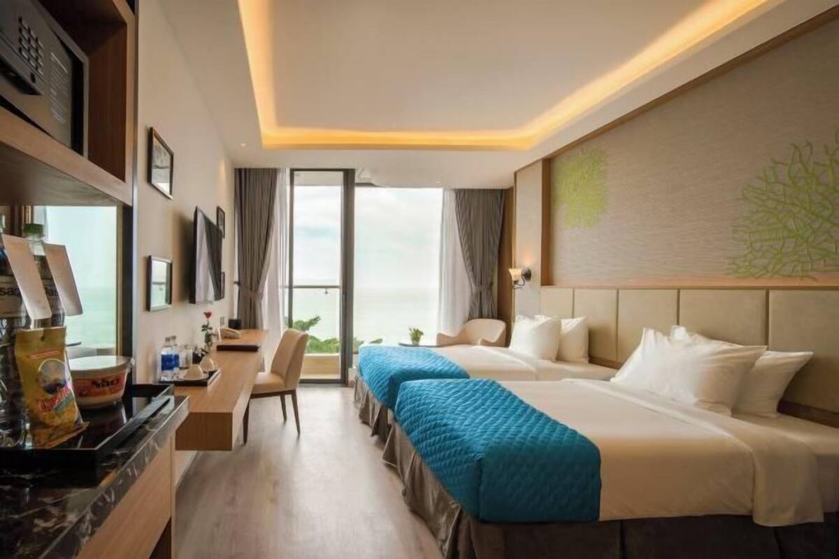 A hotel room with two beds, a desk with a chair, a large window with a view, and a wall-mounted TV.