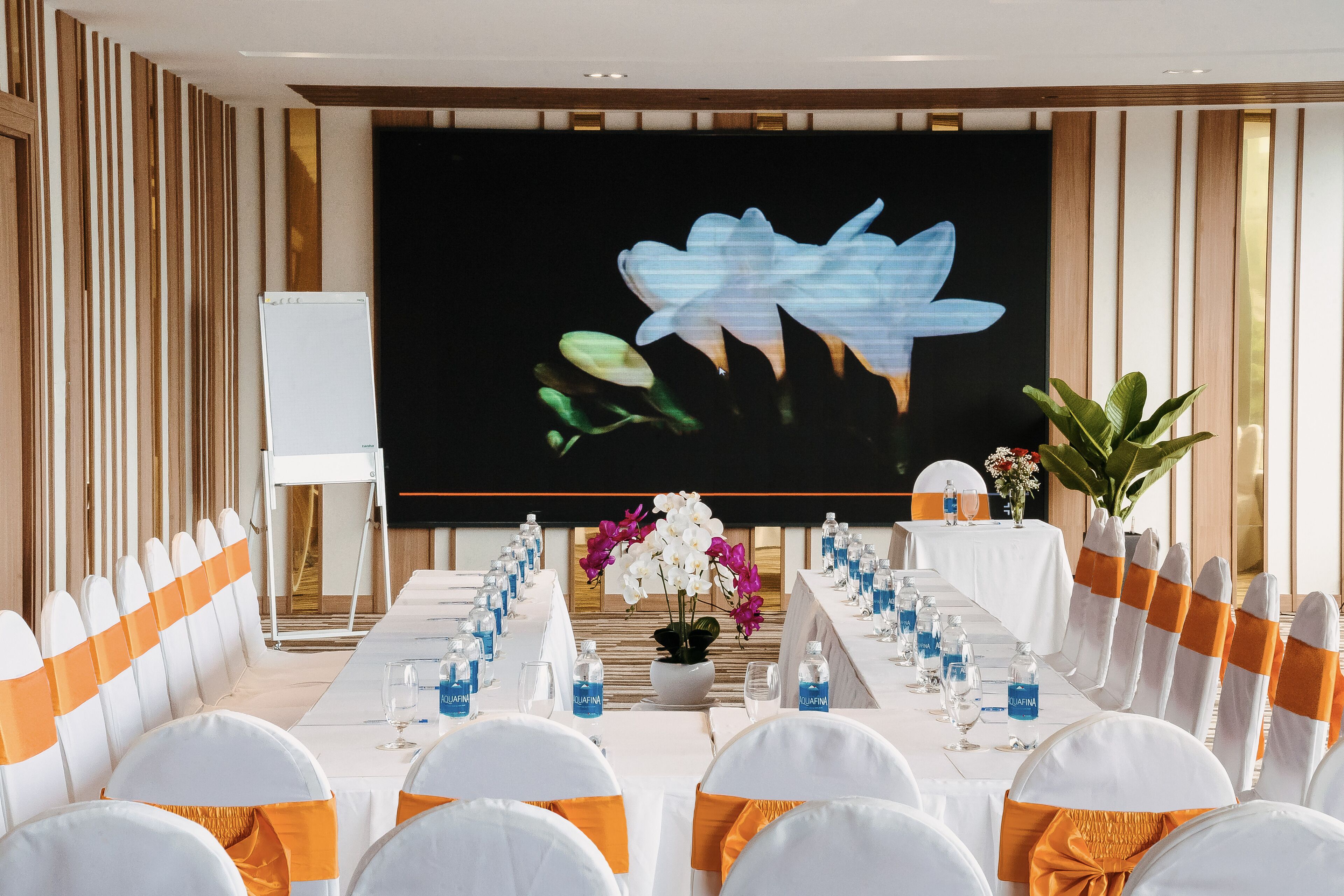 A conference room with a long table, white chairs with orange sashes, and a large screen displaying a floral image.