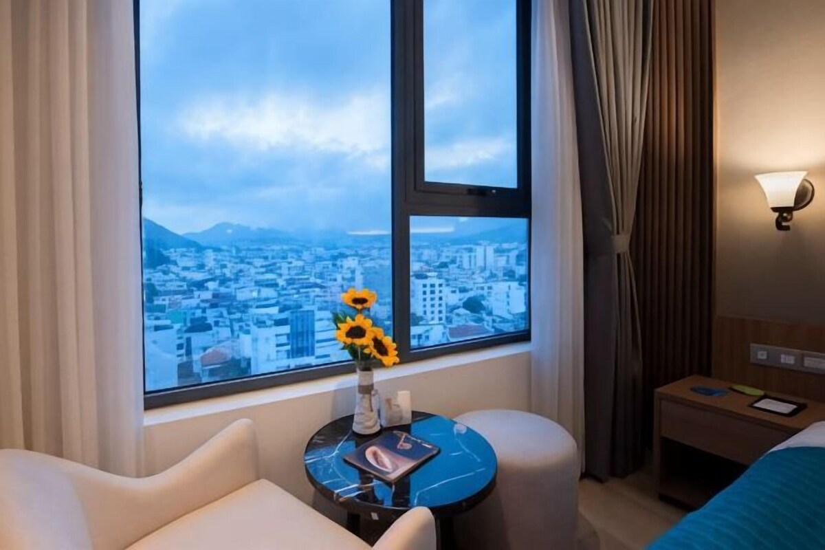 A hotel room with a large window, a vase with sunflowers, a small table, a bed, and a nightstand.