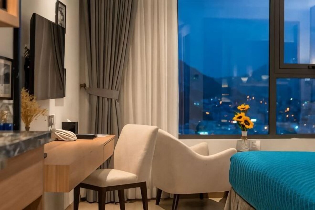 A hotel room with a desk, chair, and a bed with a teal bedspread. A window offers a city view.