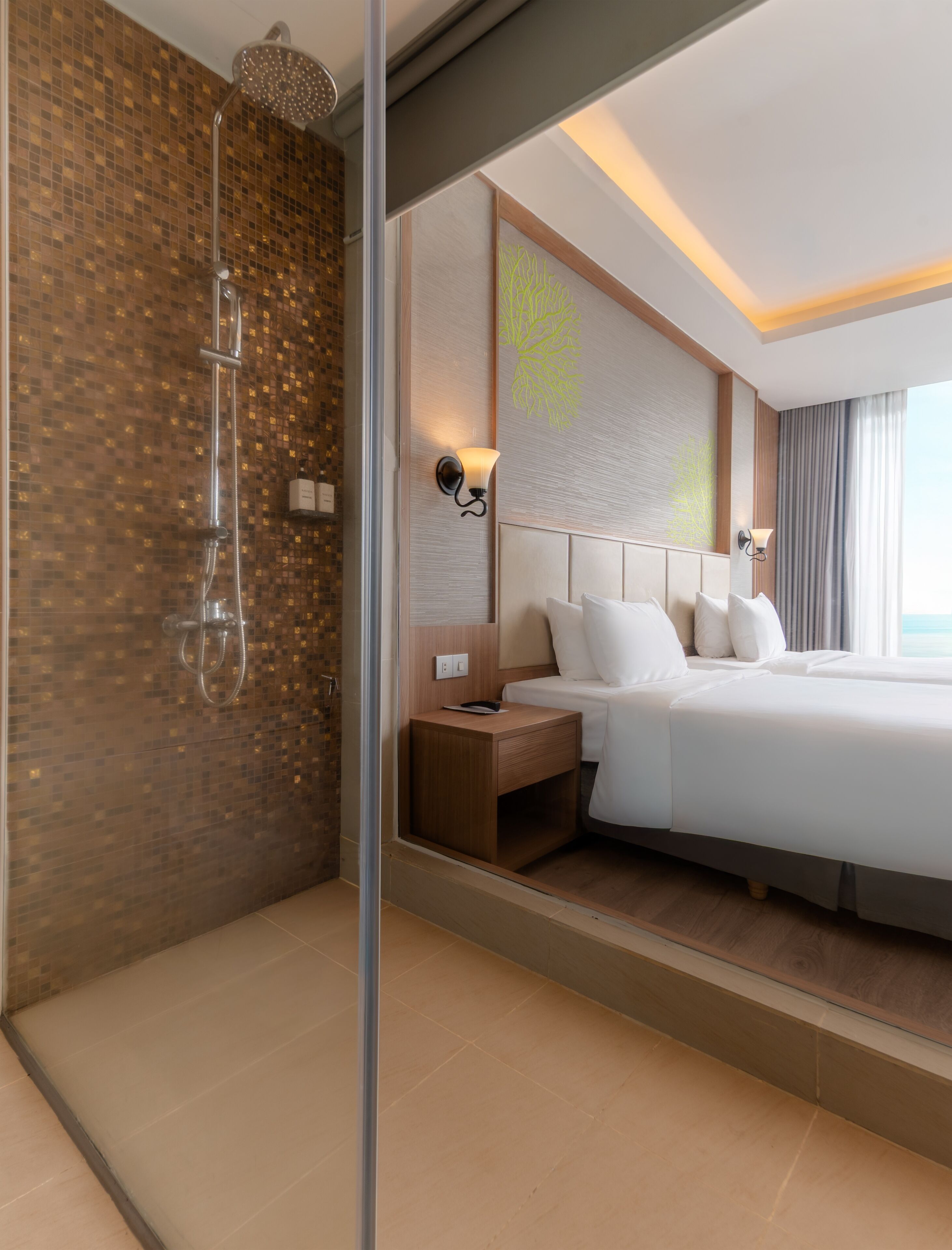 A modern hotel room with a large bed, a nightstand, a shower with a glass door, and a view of the sea through the window.