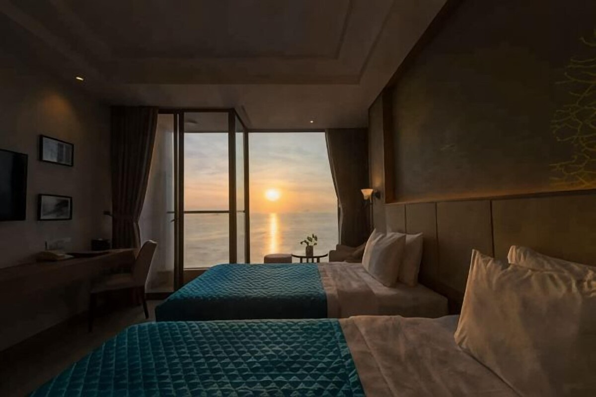 A hotel room with two beds, a desk, and a view of the sunset over the ocean.