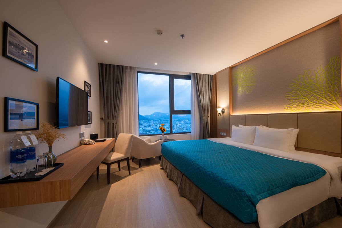 A hotel room with a large bed, a desk with a chair, a view of the city through large windows, and a mounted flat-screen TV.