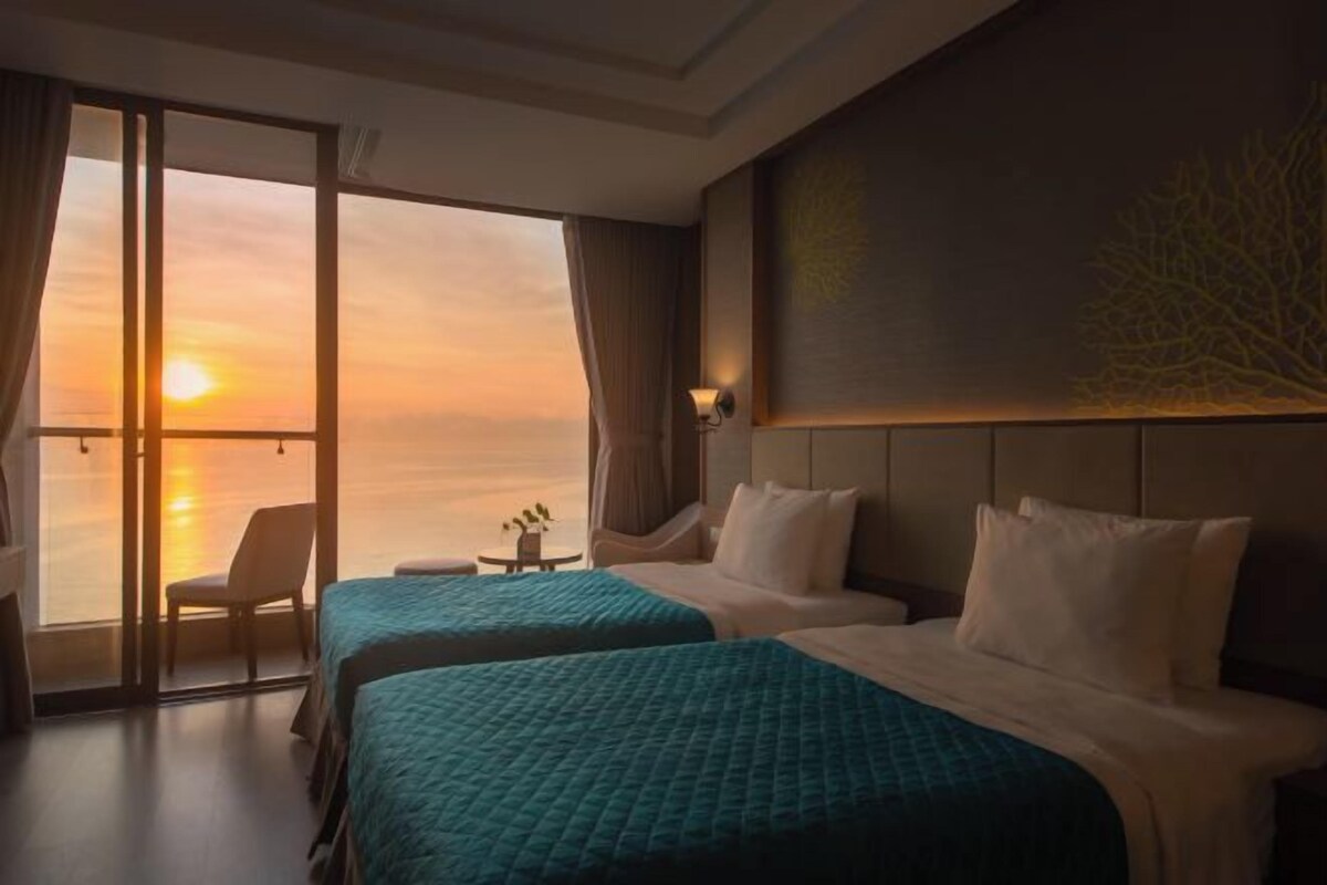 A hotel room with two beds, a view of the sunset, a chair, and a small table.