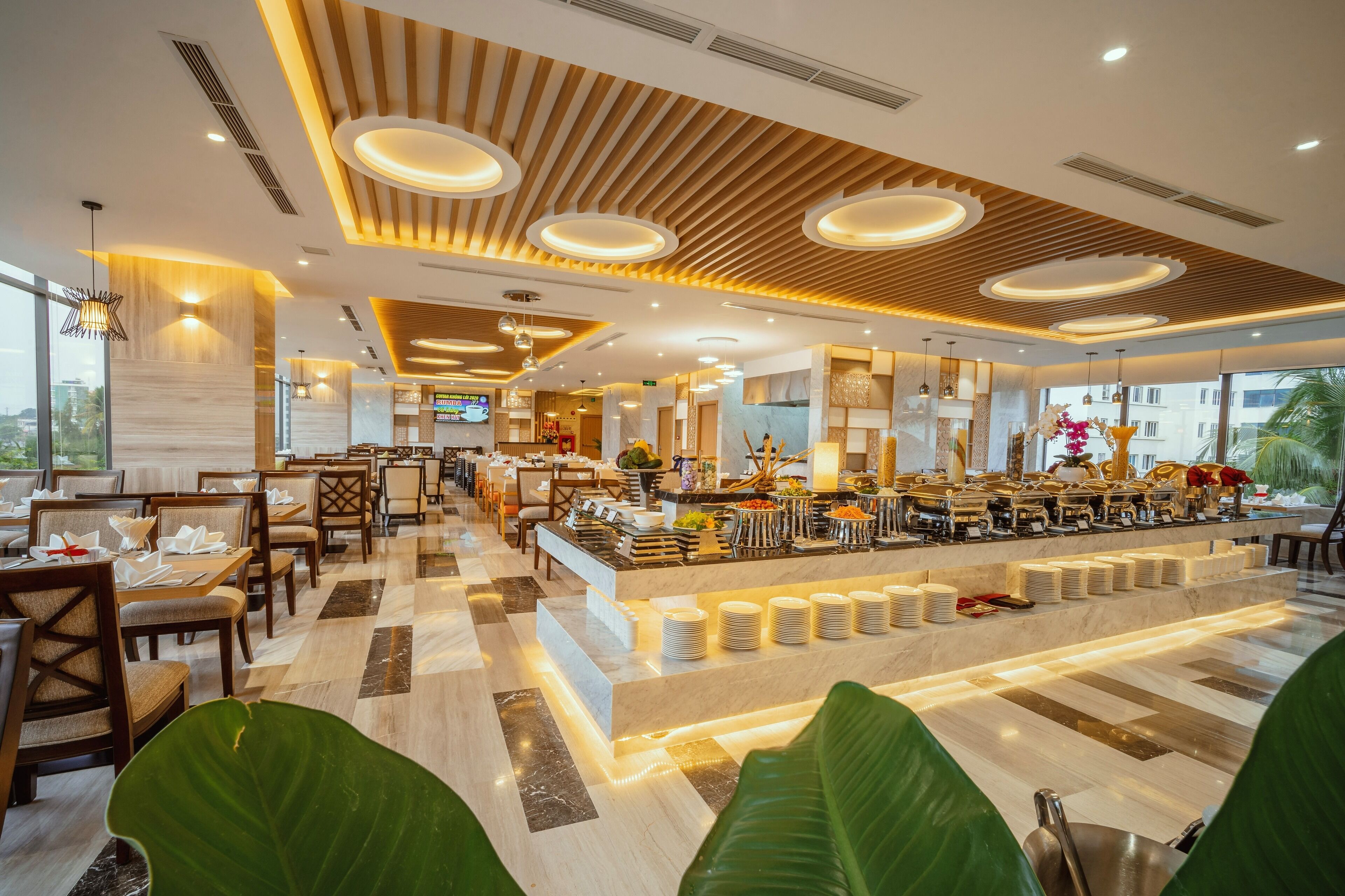 Un moderno vestíbulo de hotel con zona de buffet, arreglos de asientos y una planta grande al frente.