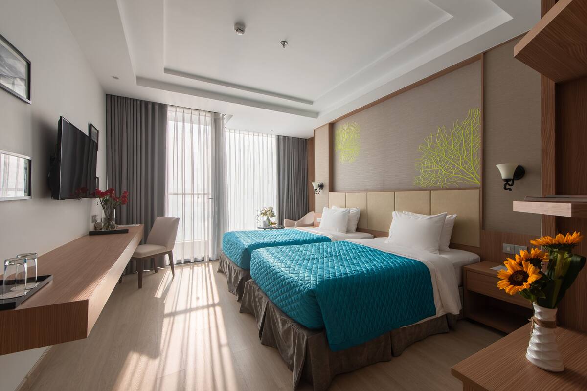 A hotel room with two beds, a desk, a chair, a TV, and a large window with curtains.