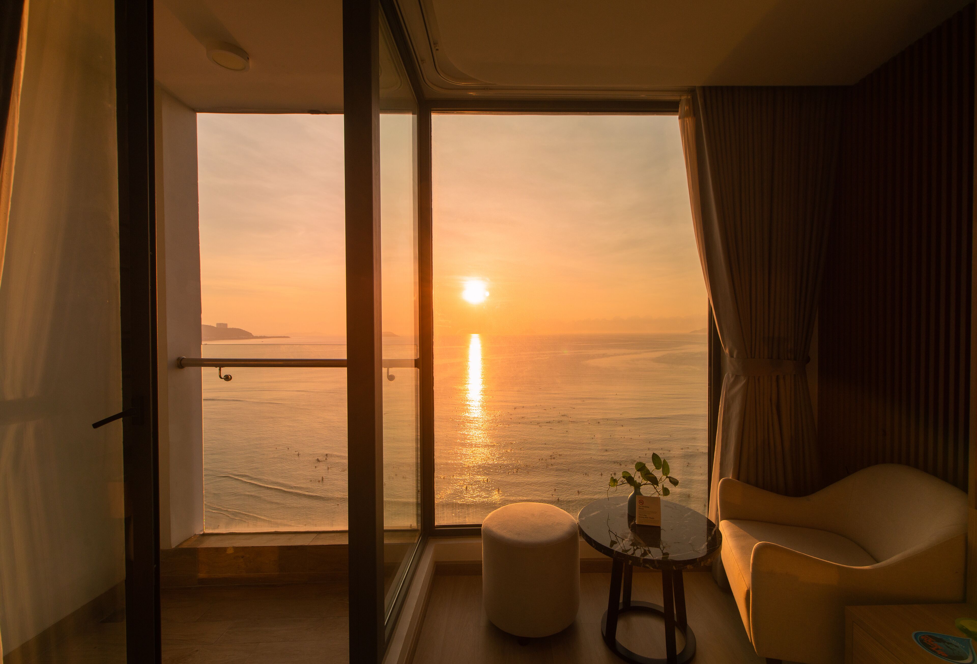 A room with a large window overlooking a sunset over the sea, a round ottoman, a small table with a plant, and a chair.