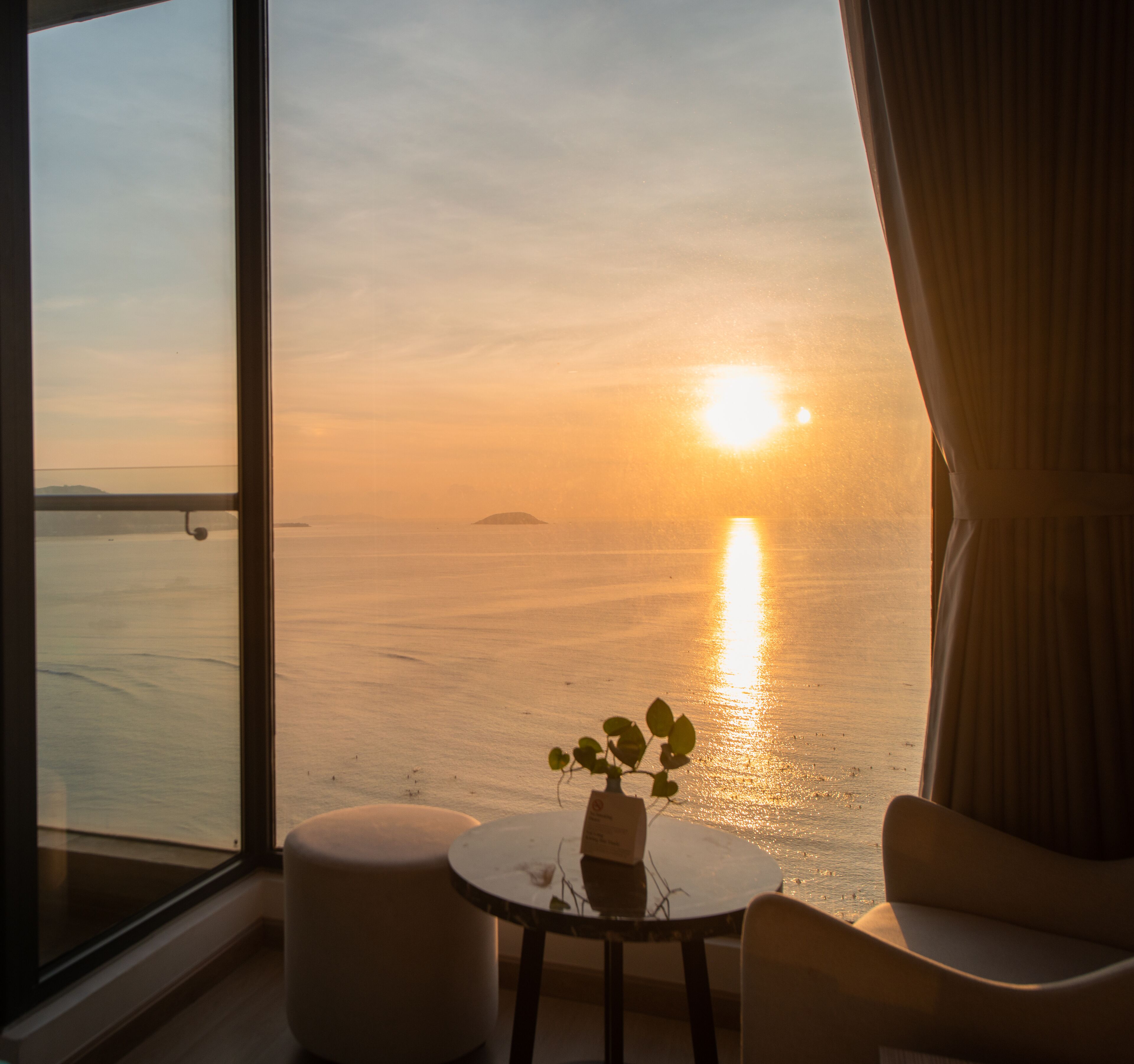 Habitación con un amplio ventanal que da a una puesta de sol sobre el agua, una pequeña mesa con una planta y una silla.