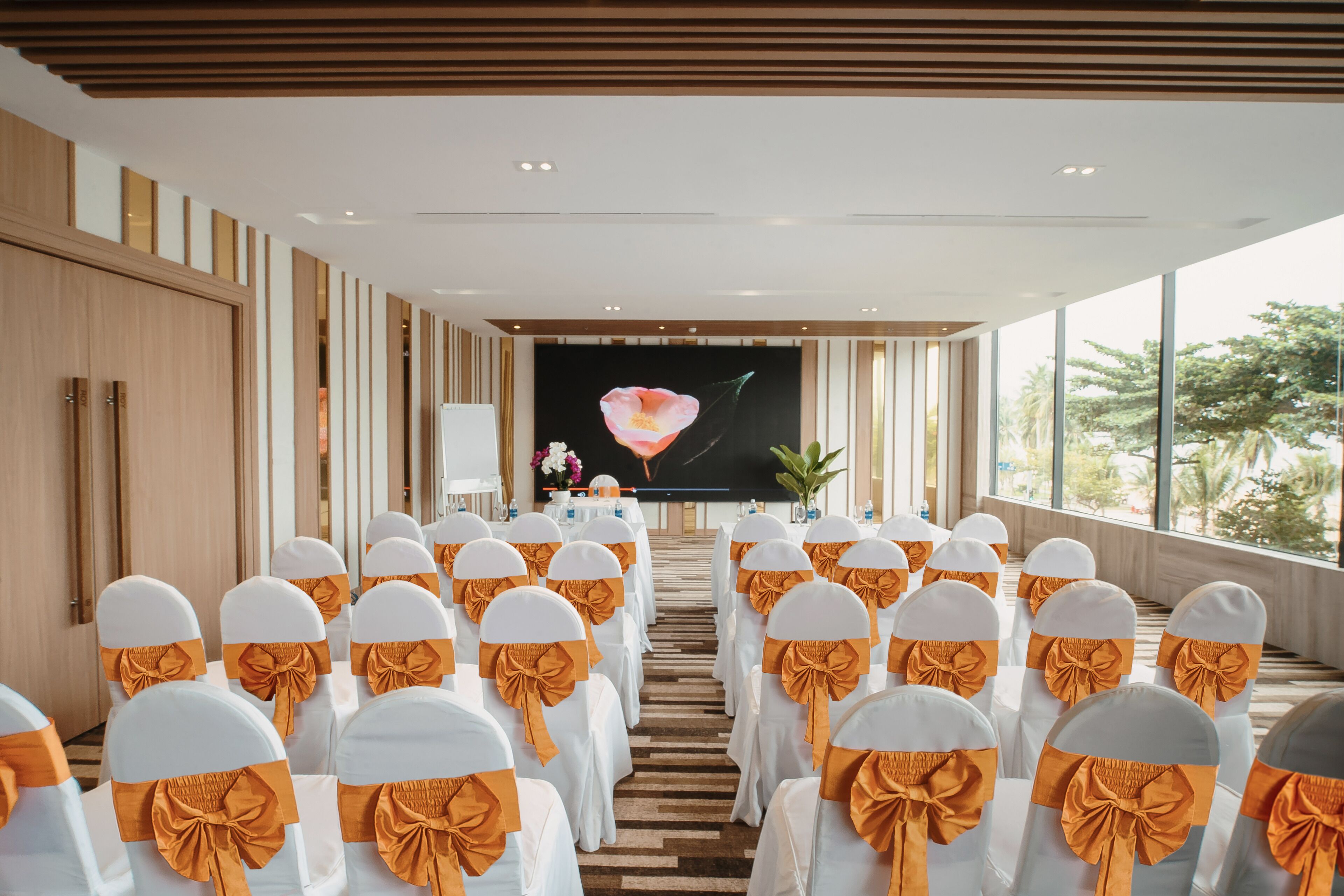 Une salle de conférence avec des rangées de chaises blanches ornées de rubans orange, un grand écran à l’avant et une vue sur les arbres à travers de grandes fenêtres.