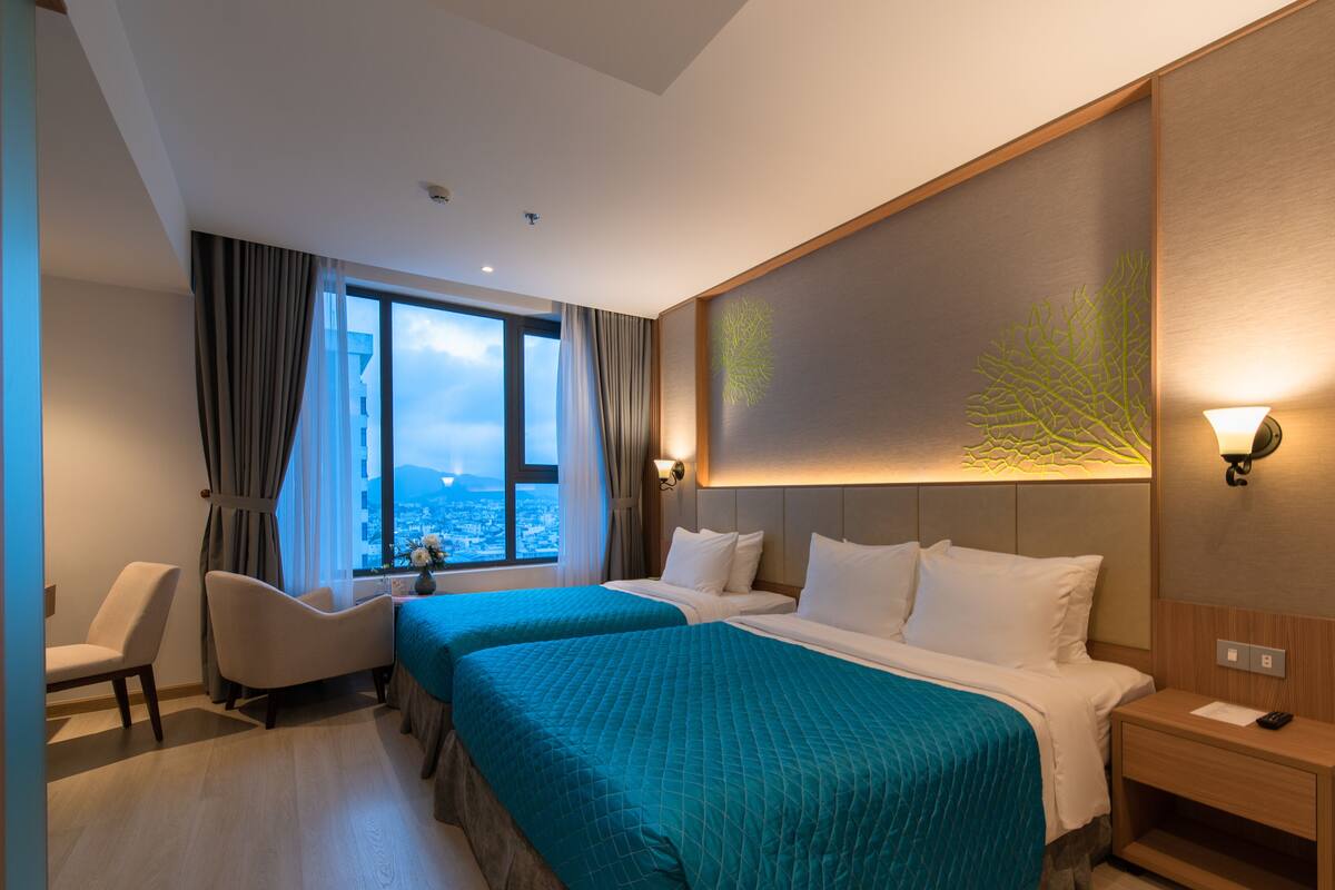 A hotel room with two beds, a desk, a chair, a window with a view, and a wall-mounted light fixture.