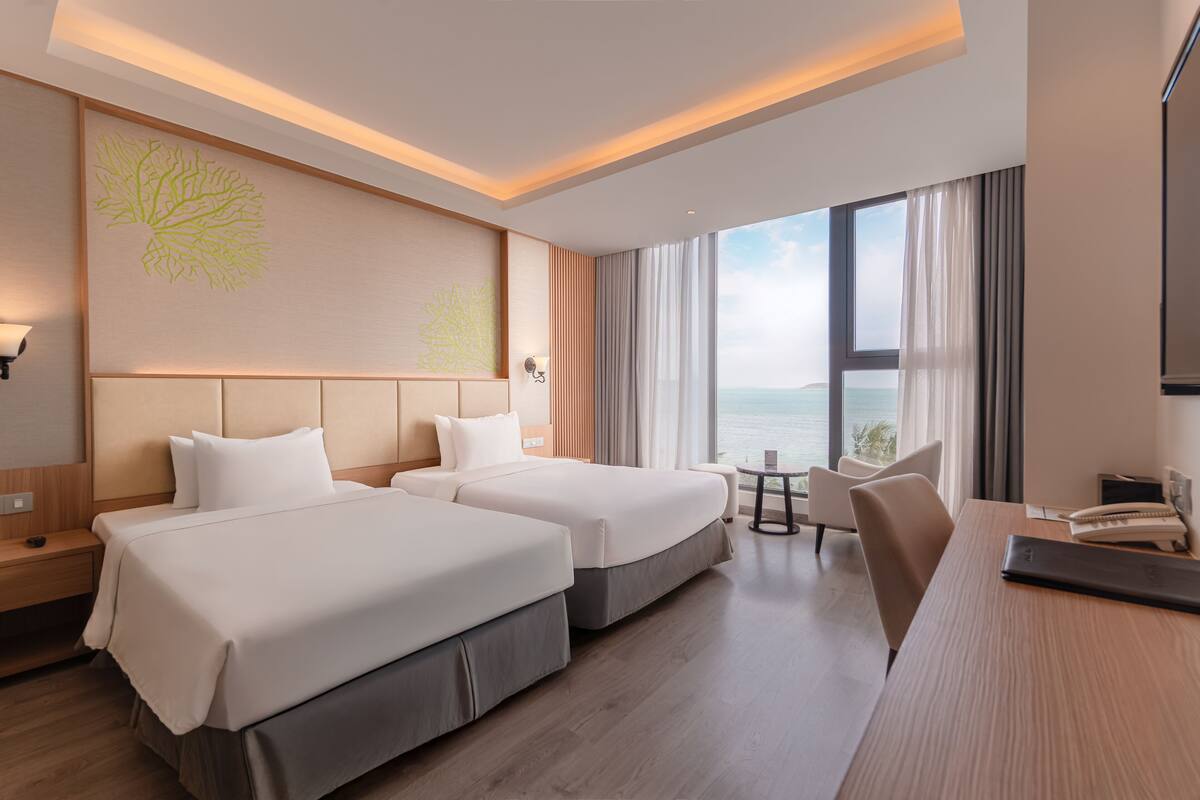A hotel room with two beds, a desk, a chair, a small table, and a large window offering a view of the sea.