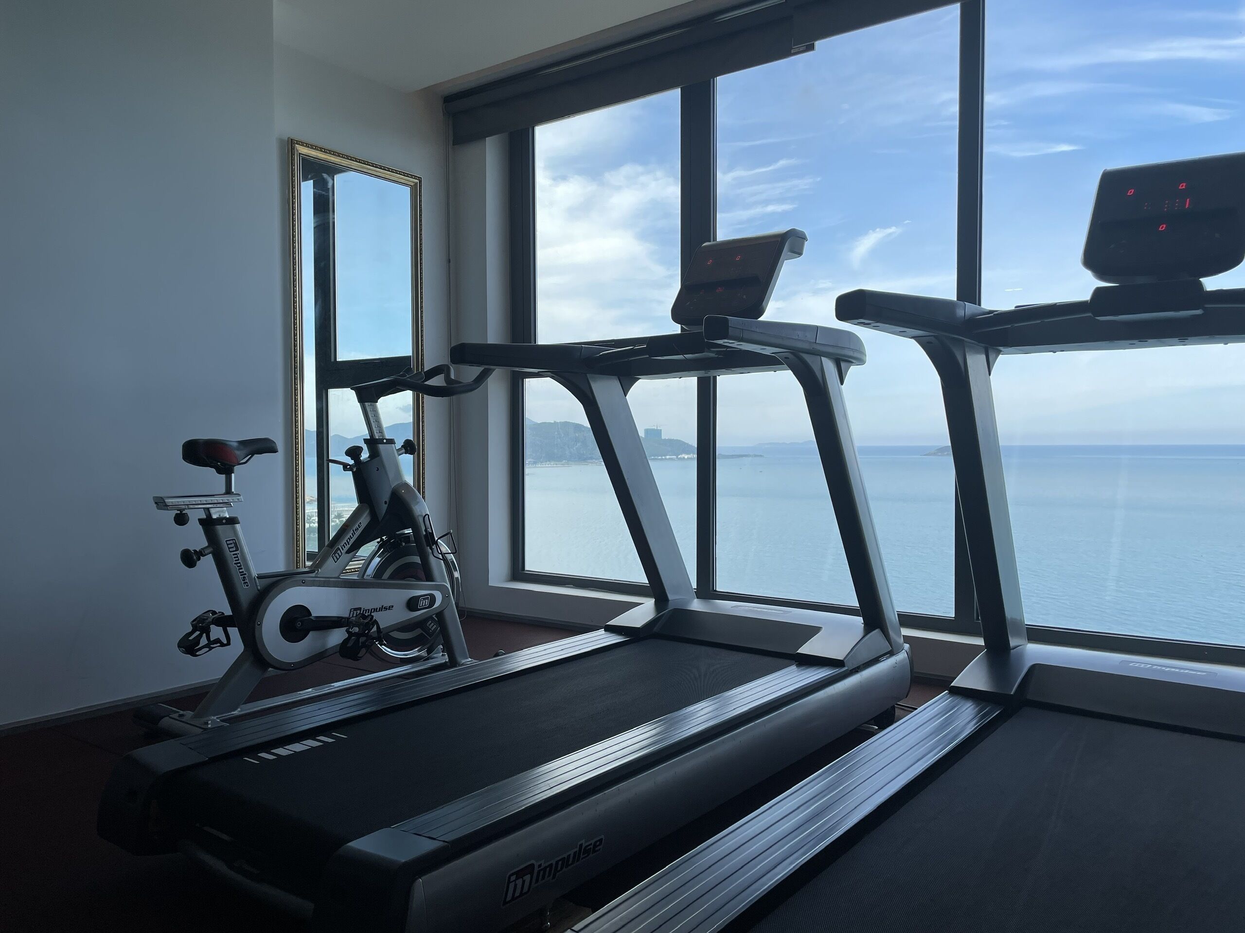 A gym with a treadmill and an exercise bike, large windows with a view of the sea.