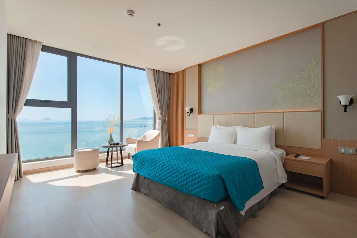 A modern hotel room with a large bed, a view of the sea, a small table with a vase, and a nightstand with a lamp.