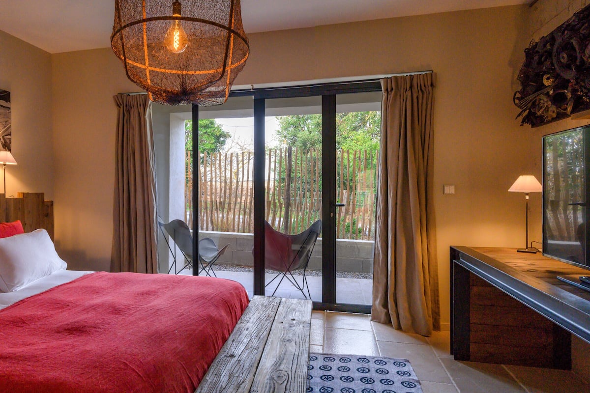 A bedroom with a large bed, a desk, a chair, and a view of a garden through a sliding door.