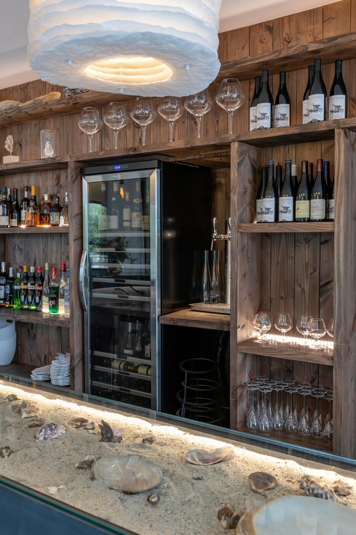 A bar area with wooden shelves displaying bottles and glasses, a refrigerator, and a sand feature with shells.