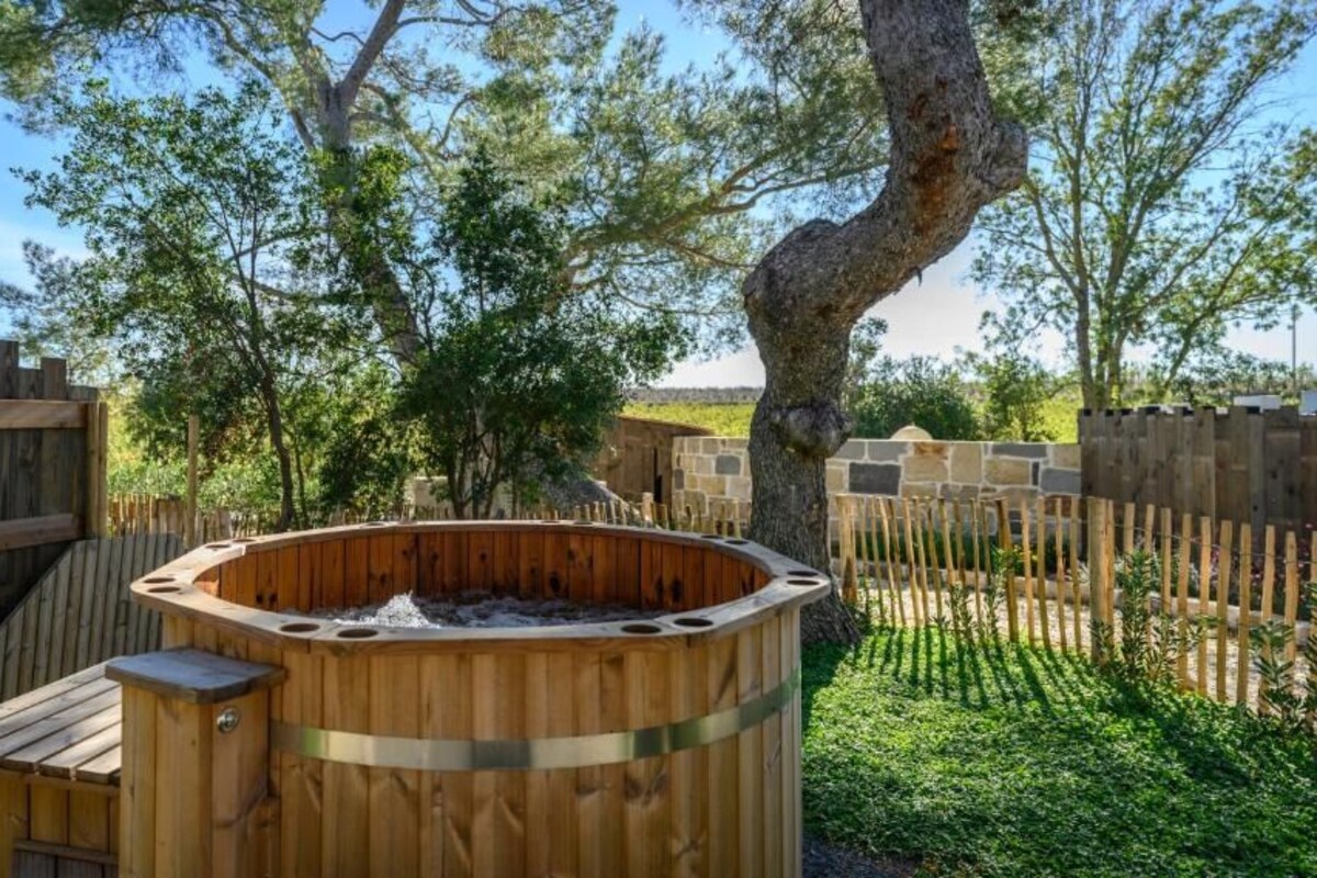 Una bañera de hidromasaje de madera rodeada por una cerca de madera, con árboles y un cielo despejado al fondo.