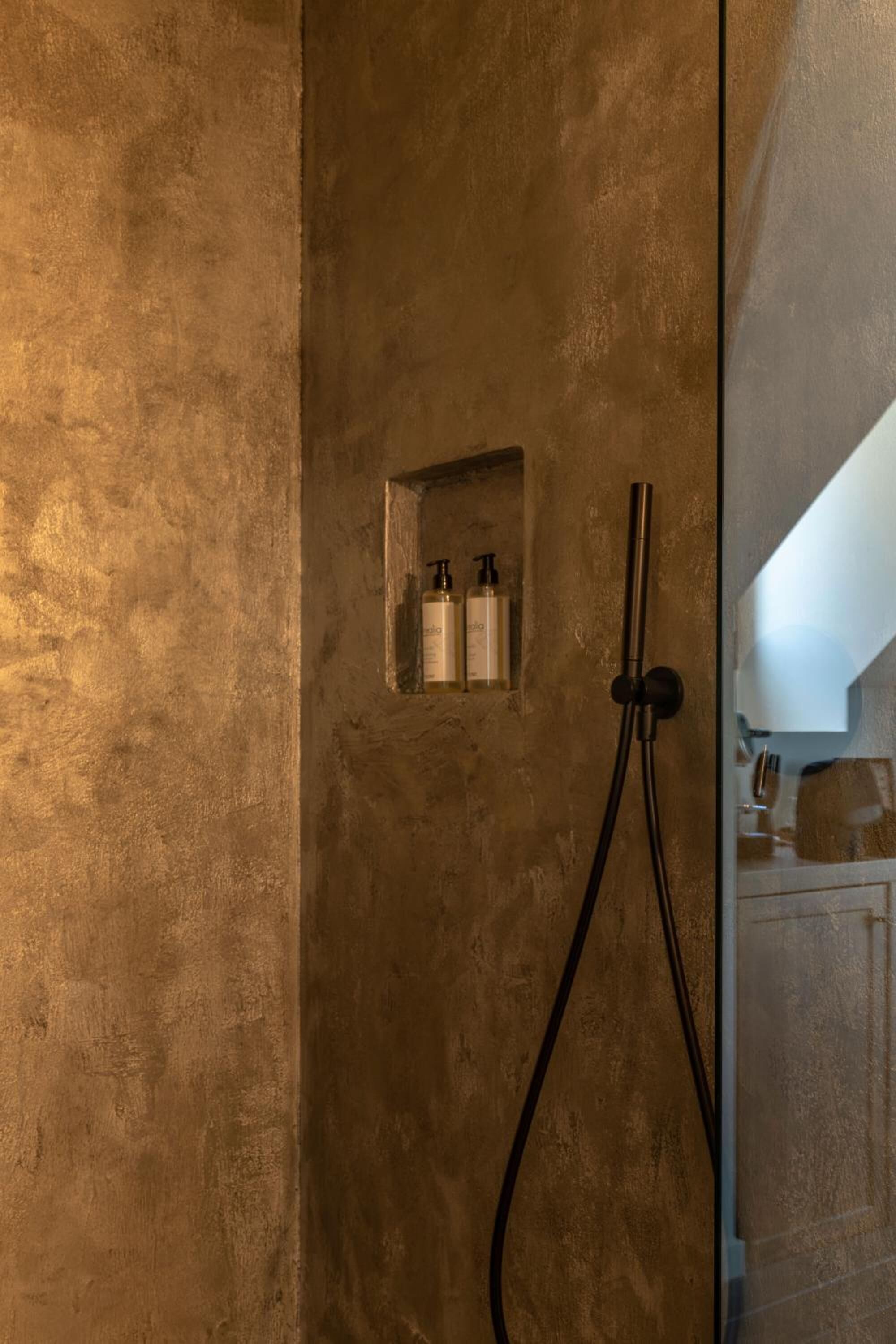 A shower area with a textured wall, a small shelf with toiletries, and a handheld showerhead.