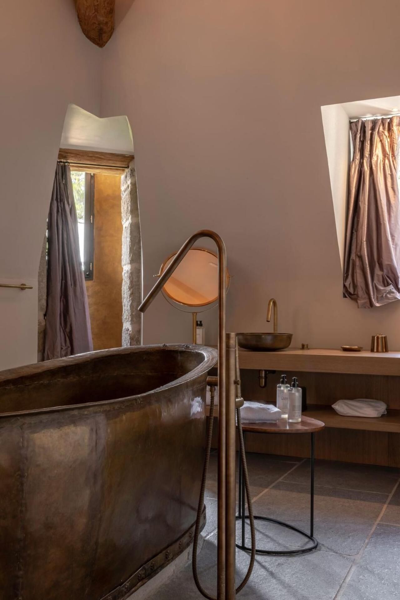 Un baño moderno con una bañera de cobre grande, un espejo redondo, un lavamanos con grifo de cobre y una ventana con cortinas.