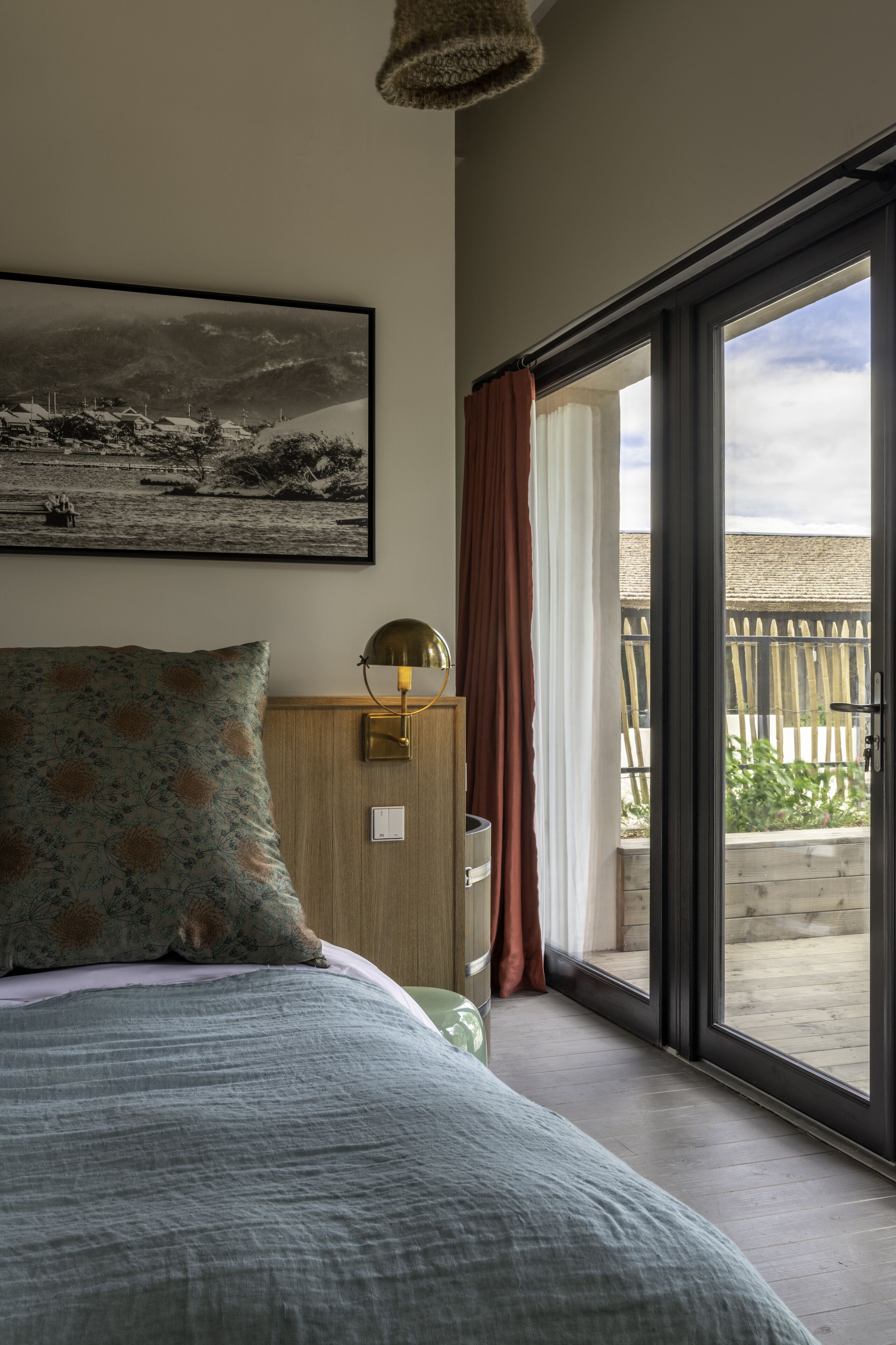 Een slaapkamer met een bed, een wandlamp, een lijst met een afbeelding en een schuifdeur naar een buitenruimte.