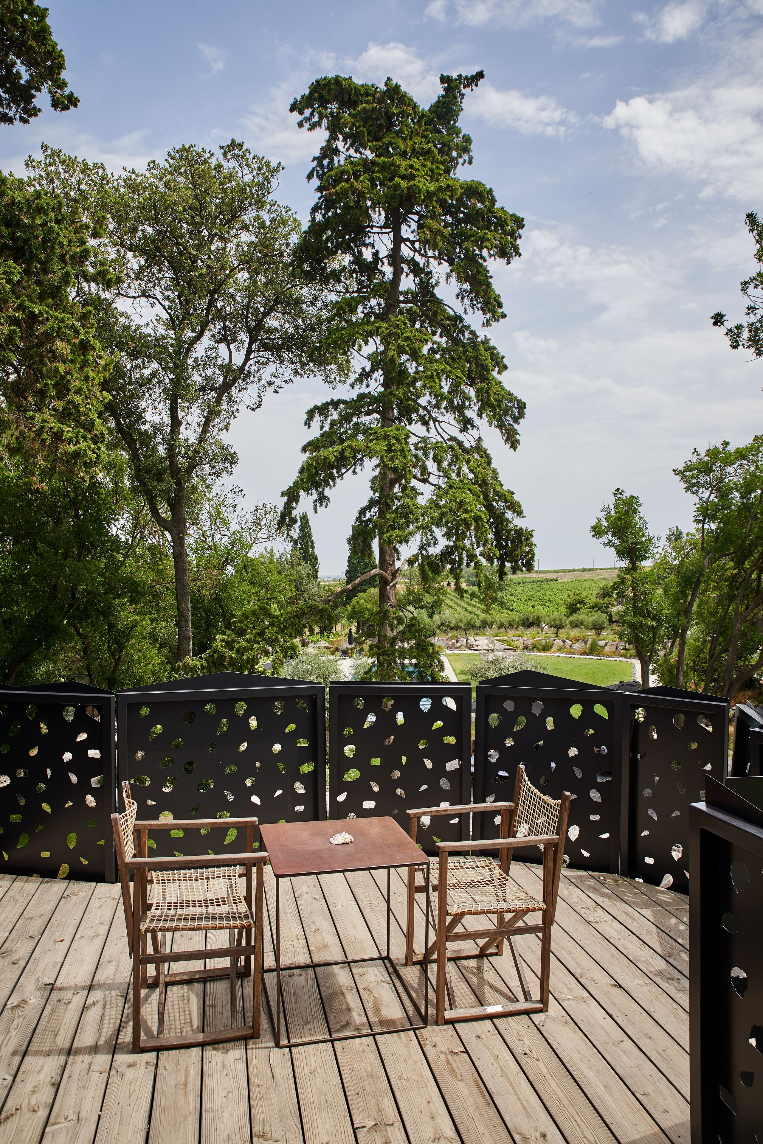 A wooden deck with two wicker chairs and a table, overlooking a lush landscape with trees and a distant view of a field.