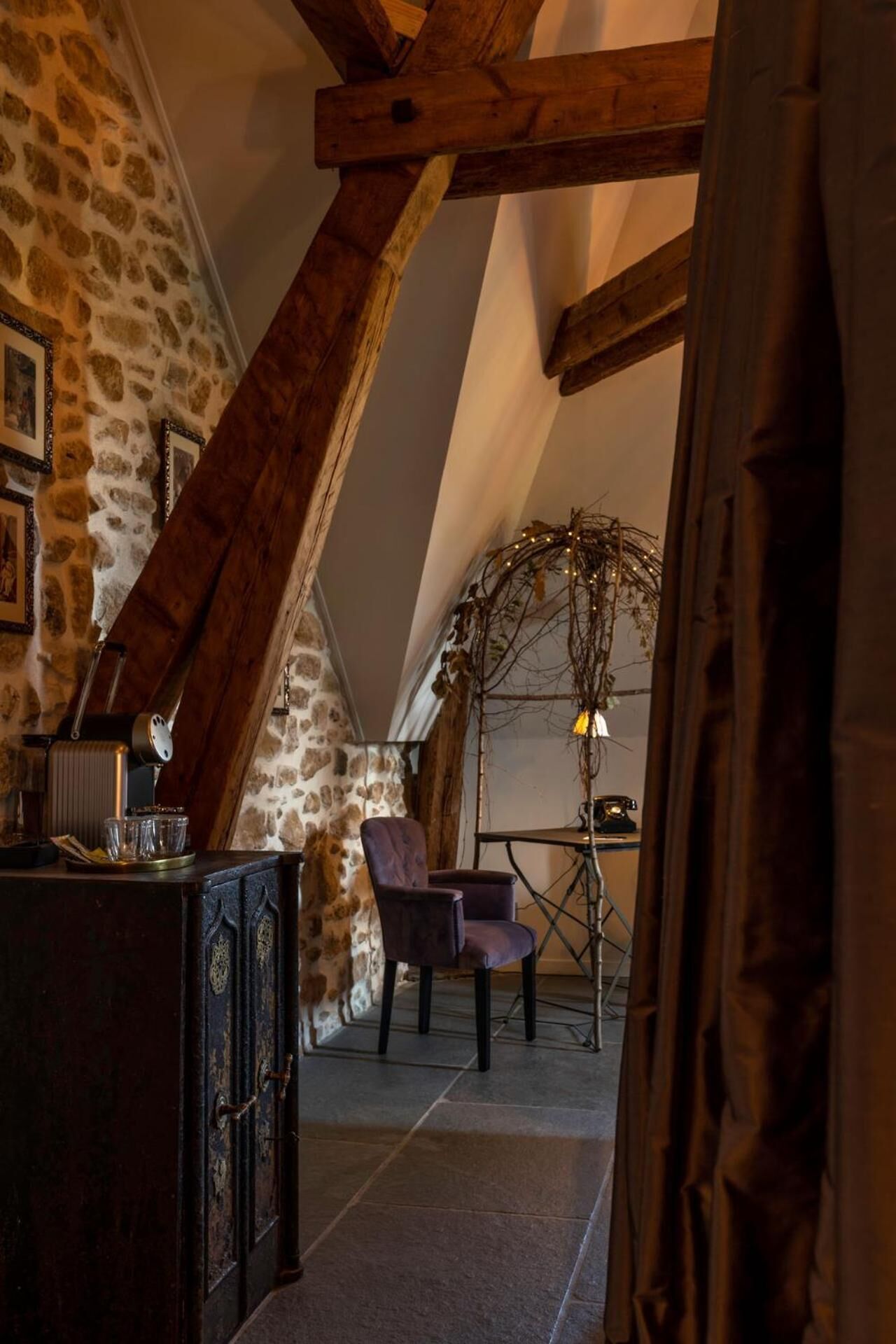 A rustic attic room with exposed wooden beams, a stone wall, and a cozy seating area with a chair and table.