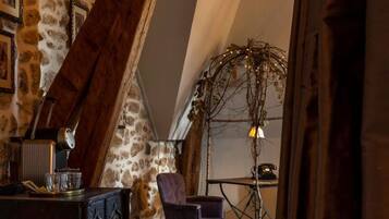 A rustic attic room with exposed wooden beams, a stone wall, and a cozy seating area with a chair and table.