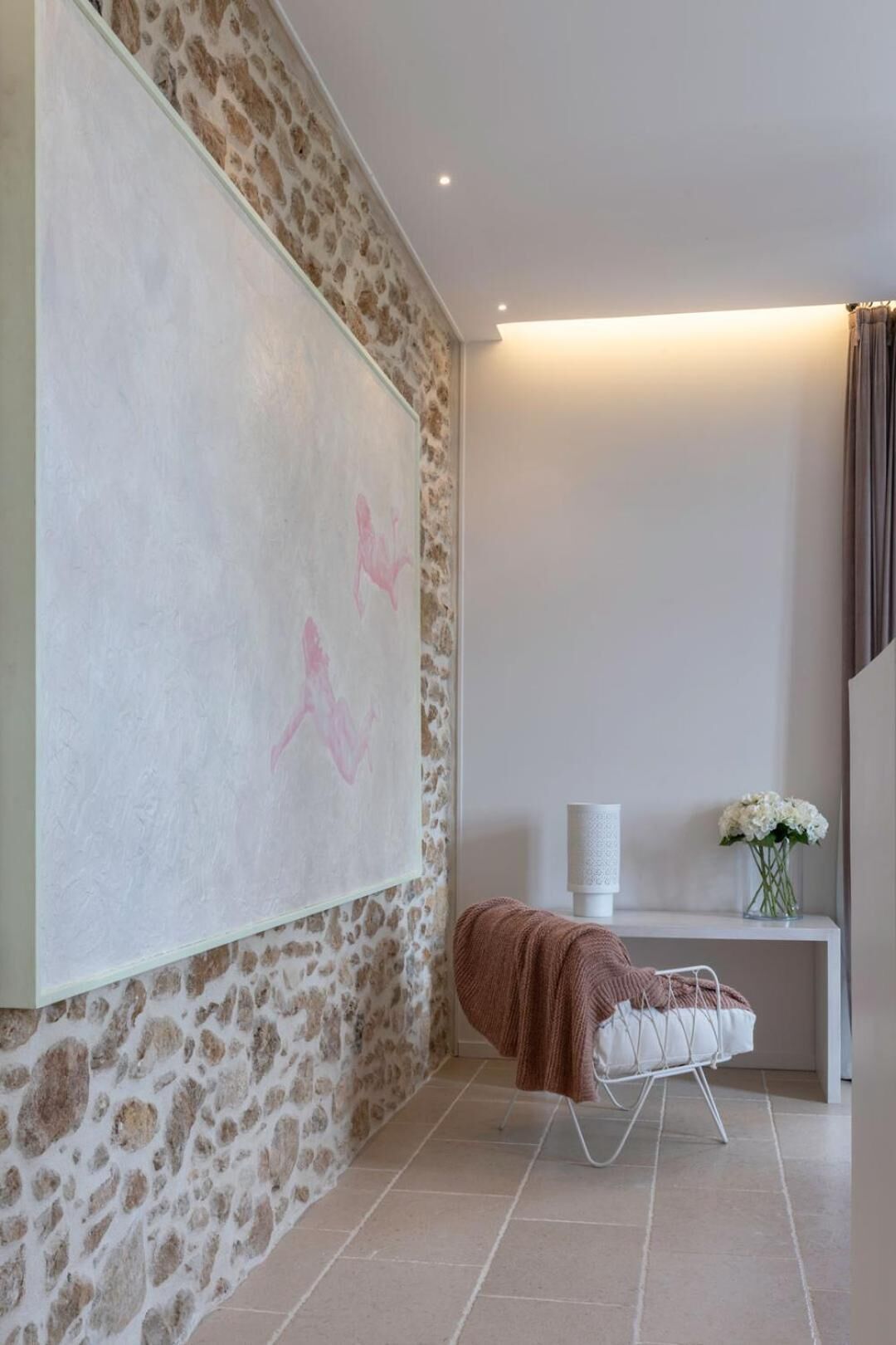 A room with a stone wall, a large abstract painting, a white console table with a vase of flowers, and a white chair with a brown blanket.